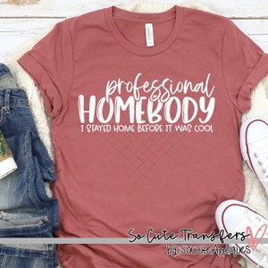 Pu&ograve; includere: Una maglietta color marrone rossiccio con il testo "professional HOMEBODY I STAYED HOME BEFORE IT WAS COOL" in bianco. La maglietta &egrave; appoggiata su una superficie di legno con un paio di scarpe da ginnastica bianche e un paio di jeans blu sullo sfondo.