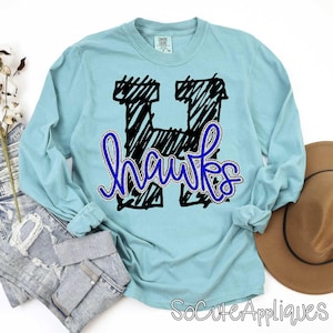 May include: A light blue long-sleeve shirt with a black and blue design. The design features a large, scribbled black letter "H" with the word "hawks" in blue script. The shirt is made of a soft material.