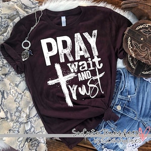 May include: A maroon t-shirt with a white graphic that says "Pray wait and trust" with a white cross in the middle of the text.