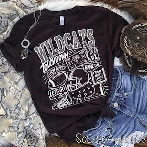 May include: A dark brown short-sleeve t-shirt featuring a white graphic print with the text WILDCATS, TOUCHDOWN, FIRST DOWN, GAME DAY, FRIDAY NIGHT LIGHTS, LETS GO, and WILDCATS, alongside illustrations of a football, helmet, and scoreboard.
