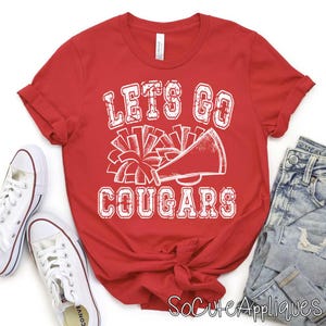 May include: A red t-shirt with the text "LET'S GO COUGARS" in white, along with a megaphone and pom-pom graphic. The shirt is paired with white sneakers and blue jeans.