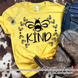 May include: Yellow t-shirt with a black bee and the word "KIND" surrounded by hearts. The shirt is on a brown and white patterned surface.