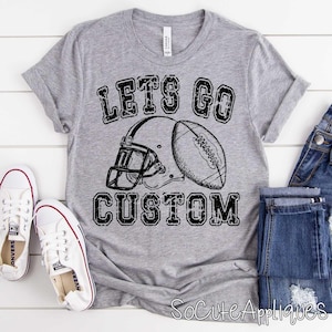 May include: A gray t-shirt with the text "LET'S GO CUSTOM" above a football helmet and football graphic. White sneakers and blue jeans are visible on a white wooden surface.