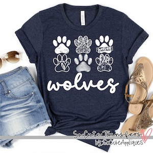 May include: A navy blue t-shirt with a white graphic of paw prints and the word "wolves". The paw prints have a distressed look and some have different patterns. The text "So Cute Transfers by: SoCuteAppliques" is at the bottom of the shirt.