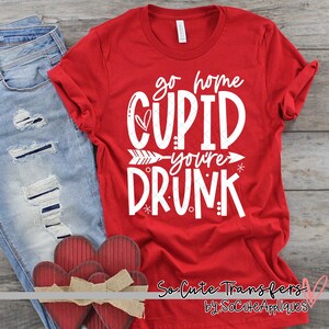 May include: Red t-shirt with white text that says "go home cupid you're drunk" with a heart and arrow graphic. The shirt is on a wooden surface with two red hearts.