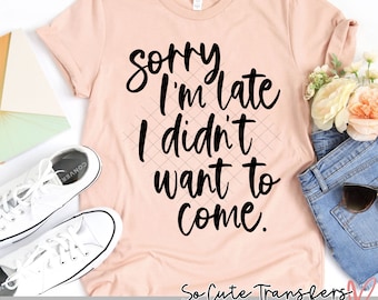 May include: A pink t-shirt with the text "Sorry I'm late I didn't want to come." in black script font. The text is on a pink t-shirt with a white background. The shirt is laying on a white surface with a pair of white sneakers and a pair of blue jeans.