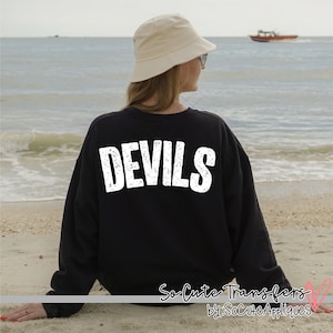 May include: Black sweatshirt with the word "DEVILS" in distressed white lettering across the back. The sweatshirt is worn by a person on a beach, with the ocean and a boat in the background. The text "So Cute Transfers by: SoCuteAppliques" is at the bottom.