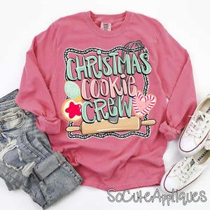 May include: A long-sleeved, dusty rose-colored shirt with the text "CHRISTMAS COOKIE CREW" in a whimsical design. The graphic includes cookie and baking-themed illustrations. The shirt is paired with jeans and white sneakers.