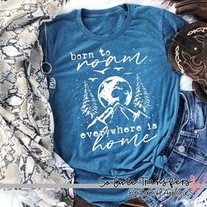 May include: A teal blue t-shirt with a white graphic design that says "born to roam everywhere is home". The graphic includes a globe, mountains, trees, and birds. The shirt is on a wooden surface with a brown and white snake skin scarf, a pair of brown cowboy boots, and a pair of blue jeans.