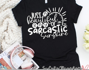 Puede incluir: Camiseta negra con el texto blanco "Just a beautiful ray of sarcastic sunshine" y un diseño de sol. La camiseta está sobre una superficie blanca con una revista y auriculares.