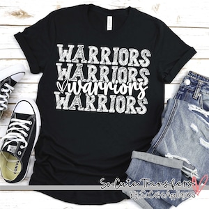 May include: Black t-shirt with the words "WARRIORS" repeated four times in white, with a heart symbol in front of the third word. The text is in a distressed, hand-drawn style. The shirt is laid flat on a wooden surface with a pair of jeans and sneakers beside it. The text "So Cute Transfers by: SoCuteAppliques" is visible in the bottom right corner.