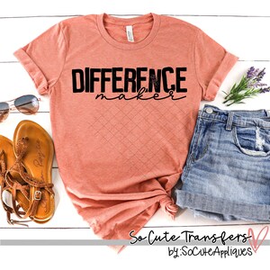 May include: A coral colored t-shirt with the text "DIFFERENCE maker" in black lettering. The shirt is laid out on a white wooden surface with a pair of brown sandals, a pair of sunglasses, and a pair of blue denim shorts.