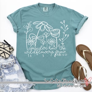 May include: A teal blue t-shirt with a white design of wildflowers and the text "Consider how the wildflowers grow... Luke 12:27".