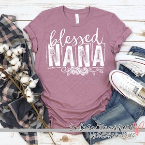 May include: A mauve t-shirt with the text "blessed NANA" in white with a distressed look. The text is framed in a white rectangle with a floral design below. The shirt is laid out on a wooden surface with a plaid shirt, cotton balls, blue jeans, and white sneakers.
