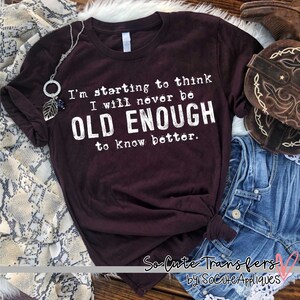 May include: A maroon t-shirt with white text that reads "I'm starting to think I will never be OLD ENOUGH to know better." The shirt is laying on a wooden surface with a pair of jeans and a brown leather boot.
