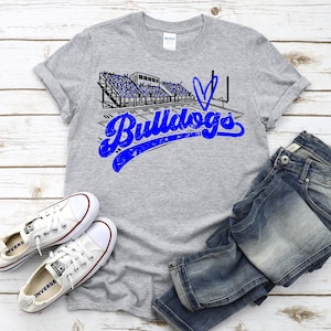 May include: A gray t-shirt with a blue and black graphic of a football stadium and the word "Bulldogs" in blue script. The shirt is paired with blue jeans and white Converse sneakers.