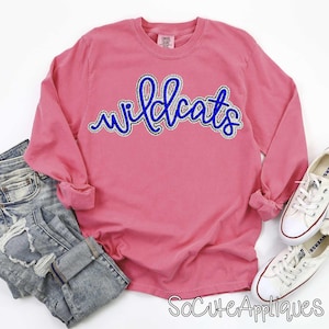 May include: A pink long-sleeve shirt with the word "wildcats" in blue script with a white outline. The shirt is paired with blue jeans and white sneakers. The image also includes the text "SoCuteAppliques".