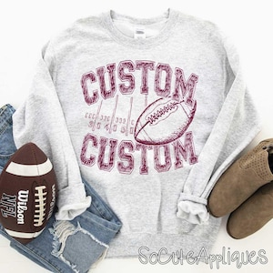 May include: A light gray sweatshirt with the word "CUSTOM" in maroon, along with a football graphic. A football and a pair of boots are also in the image. The sweatshirt design is reminiscent of a football field.