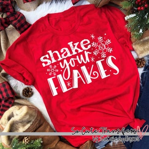 May include: Red t-shirt with white text that says "Shake Your Flakes" with snowflake designs. The shirt is on a white surface with brown and black plaid fabric and a pine cone.