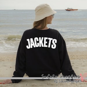 May include: Black sweatshirt with the word "JACKETS" printed in white on the back. The sweatshirt is worn by a person wearing a beige bucket hat and sitting on a sandy beach.