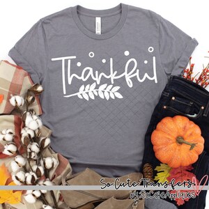 May include: Gray t-shirt with the word "Thankful" in white script with a white leafy branch underneath. The shirt is laying on a table with a plaid blanket, cotton bolls, jeans, a pumpkin, and fall leaves.