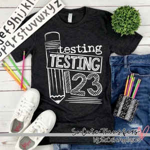 May include: A dark grey t-shirt with a white graphic of a pencil and the text "testing TESTING 123". The text is in a chalkboard style font. The shirt is laid out on a wooden surface with a pair of white sneakers, a pair of blue jeans, and a pencil holder filled with colorful pencils.