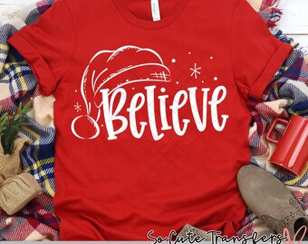 May include: Red t-shirt with a white design of a Santa hat and the word "Believe".
