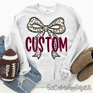 May include: A light gray sweatshirt with a large bow design featuring a leopard print pattern. The word "CUSTOM" is printed in a burgundy color across the bow. A football and a pair of boots are visible.
