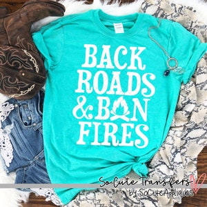 May include: Teal blue t-shirt with white text that reads "Back Roads & Bon Fires". The shirt is styled with a knot in the front and is laid on a white surface with a pair of brown cowboy boots and a denim skirt.