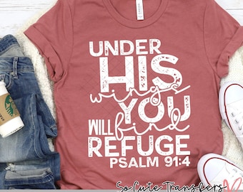 May include: A maroon t-shirt with a white distressed graphic that reads "Under His Wings You Will Find Refuge Psalm 91:4". The graphic is surrounded by small white hearts. The shirt is laid flat on a white surface with a pair of blue jeans and a coffee cup.