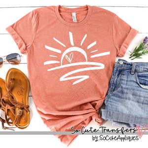 May include: A coral colored t-shirt with a white graphic of a sun with a heart in the middle. The graphic is a simple line drawing style. The t-shirt is laying on a white surface with a pair of sandals and a pair of blue jeans.