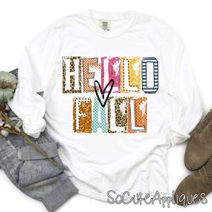 Può includere: Maglietta bianca a maniche lunghe con una grafica colorata "Hello Fall" che include un design a forma di cuore. La grafica è composta da diversi motivi, tra cui stampa leopardata, pois e strisce.
