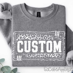 May include: A heather gray long-sleeve sweatshirt featuring a white rectangular graphic print with a distressed border. The design includes a leopard print pattern at the top and bottom, with the word CUSTOM printed in large white block letters.