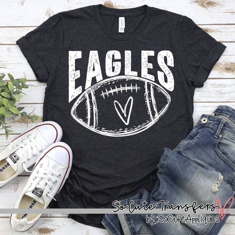 Puede incluir: Una camiseta gris oscura con un gr&aacute;fico blanco de un bal&oacute;n de f&uacute;tbol con un coraz&oacute;n en su interior y la palabra "EAGLES" encima. La camiseta est&aacute; colocada sobre una superficie de madera con un par de zapatillas blancas y un par de pantalones vaqueros azules.