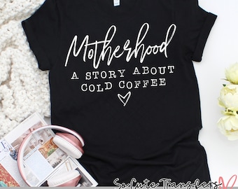 May include: Black t-shirt with white text that reads "Motherhood A Story About Cold Coffee" with a heart symbol. The shirt is on a white surface with a magazine and headphones.