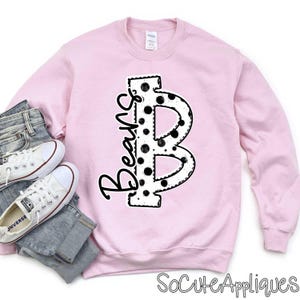 May include: A light pink sweatshirt with a large black and white polka-dot letter "B" and the word "Beans" in a script font. The sweatshirt is paired with blue jeans and white canvas sneakers.