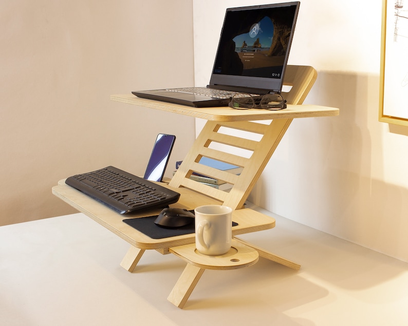 May include: A wooden standing desk with a laptop, keyboard, mouse, phone, and a cup on it. The desk is made of light wood and has a simple design.