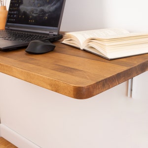 Puede incluir: Una estanter&iacute;a de escritorio de madera con un borde redondeado, montada en una pared blanca. Un ordenador port&aacute;til, un rat&oacute;n inal&aacute;mbrico y un libro est&aacute;n en la estanter&iacute;a.