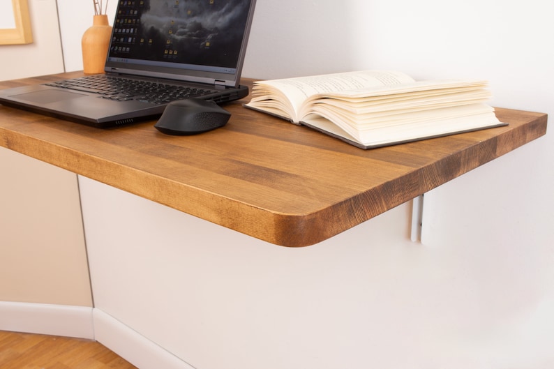 Puede incluir: Una estanter&iacute;a de escritorio de madera con un borde redondeado, montada en una pared blanca. Un ordenador port&aacute;til, un rat&oacute;n inal&aacute;mbrico y un libro est&aacute;n en la estanter&iacute;a.