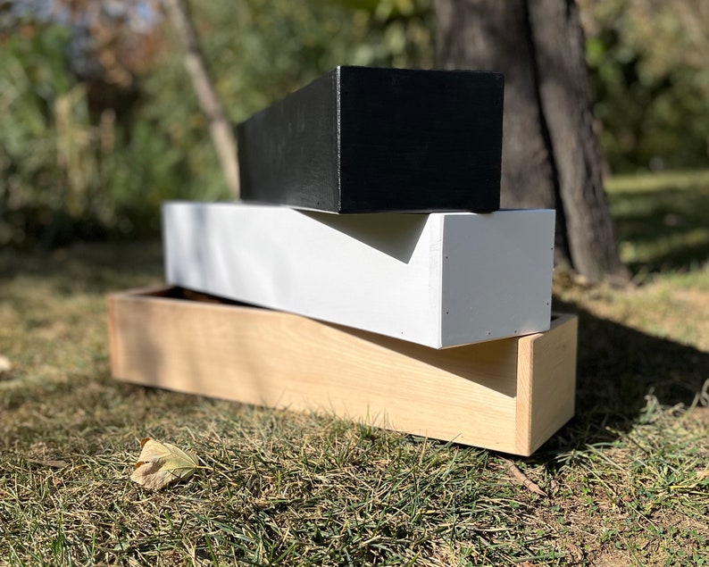 May include: Three wooden planter boxes stacked on top of each other. The top box is black, the middle box is white, and the bottom box is natural wood.