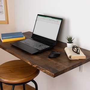Pode incluir: Uma prateleira flutuante de madeira castanho escura com um laptop, um rato sem fios, um livro, uma suculenta em vaso e uns &oacute;culos. A prateleira est&aacute; montada numa parede branca.