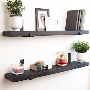 May include: Two black wooden shelves with metal brackets mounted on a white wall. The top shelf has a potted plant, a framed photo, and a few books. The bottom shelf has a candle, a snow globe, a small box, and a potted plant.