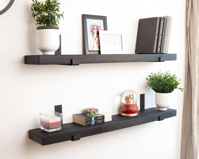 May include: Two black wooden shelves with metal brackets mounted on a white wall. The top shelf has a potted plant, a framed photo, and a few books. The bottom shelf has a candle, a snow globe, a small box, and a potted plant.