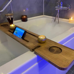Puede incluir: Una bandeja de baño de madera con un soporte para teléfono, dos portavelas y un portavasos. La bandeja está en una bañera blanca con burbujas y una luz azul.