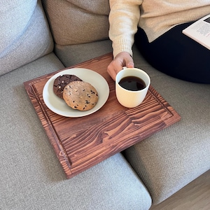 May include: A wooden tray with a brown stain sits on a grey sofa. The tray holds a white ceramic mug filled with coffee and a white plate with three chocolate chip cookies.