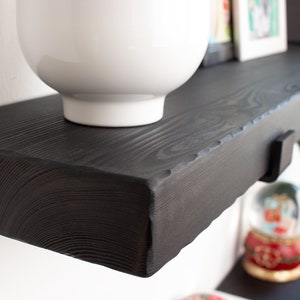 May include: A black wooden shelf with a white bowl on top. The shelf has a rough, rustic edge and is mounted to a white wall.