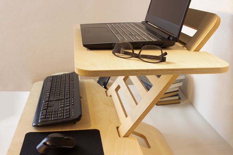 May include: A light wood standing desk with a laptop, keyboard, mouse, and glasses on top. The desk has a built-in shelf for storage.