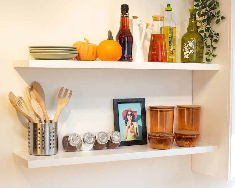 May include: White shelves with various kitchen items, including a framed photo, glass jars, wooden spoons, and a plate. There are also two pumpkins and several bottles of oil and vinegar.