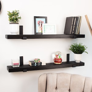 May include: Two black floating shelves with black metal brackets, mounted on a white wall. The top shelf has a potted plant, a framed photo, and a stack of books. The bottom shelf has a candle, a snow globe, and a stack of books.