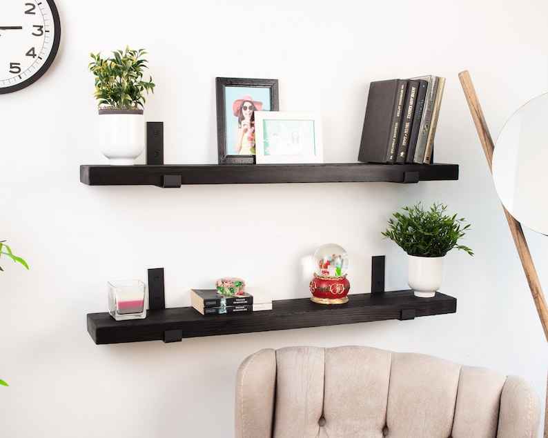 May include: Two black floating shelves with black metal brackets, mounted on a white wall. The top shelf has a potted plant, a framed photo, and a stack of books. The bottom shelf has a candle, a snow globe, and a stack of books.